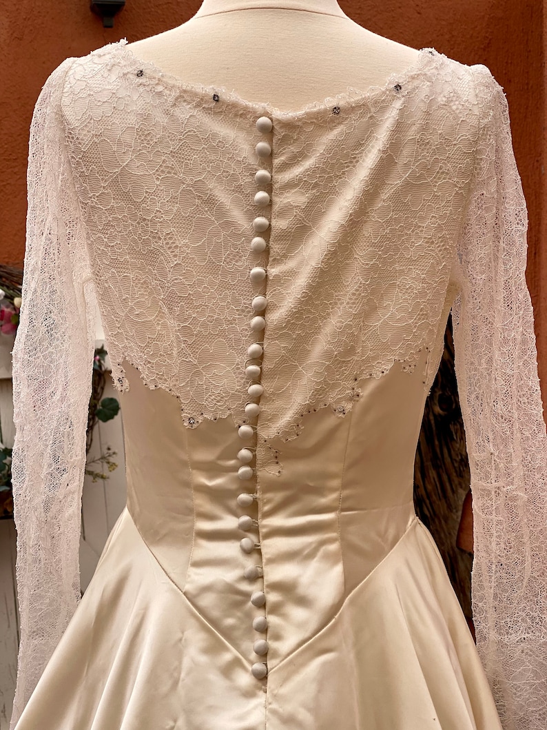 May include: A white satin wedding dress with a lace overlay and a row of buttons down the back.