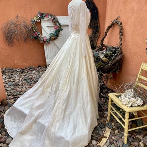 May include: A vintage ivory satin wedding dress with a lace overlay and a long train. The dress is on a mannequin and is displayed against a backdrop of a stone wall and greenery.