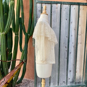 May include: A cream-coloured lace top with a ruffled collar and short sleeves. The top is displayed on a mannequin against a wooden background.