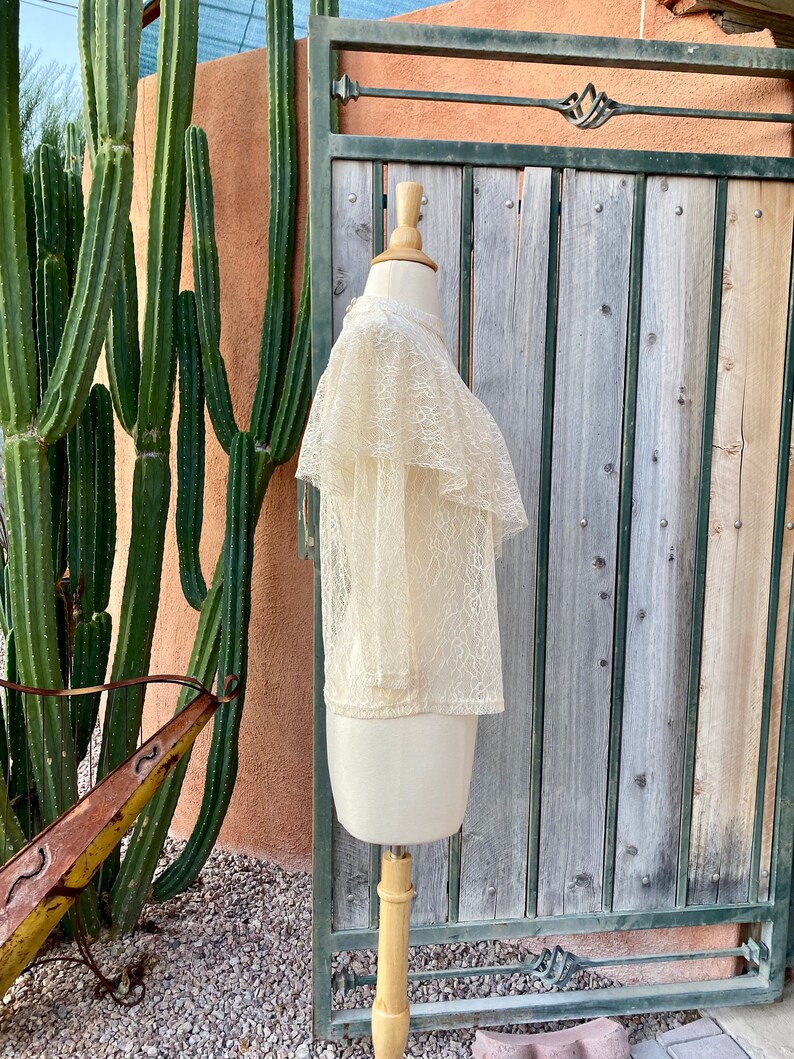 May include: A cream-coloured lace top with a ruffled collar and short sleeves. The top is displayed on a mannequin against a wooden background.