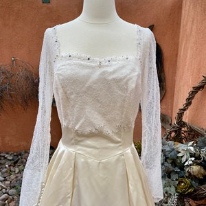 May include: A white satin wedding dress with a lace bodice and long sleeves. The bodice is adorned with small, clear rhinestones.