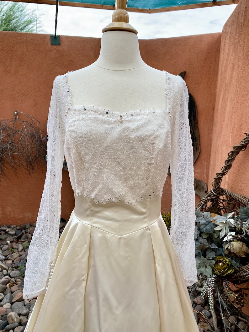 May include: A white satin wedding dress with a lace bodice and long sleeves. The bodice is adorned with small, clear rhinestones.