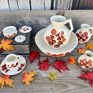 May include: A set of vintage white ceramic bathroom accessories with a floral pattern in shades of orange and brown. The set includes a pitcher, a bowl, a soap dish, a lidded jar, and a small vase. The items are arranged on a wooden surface with fall leaves scattered around them.