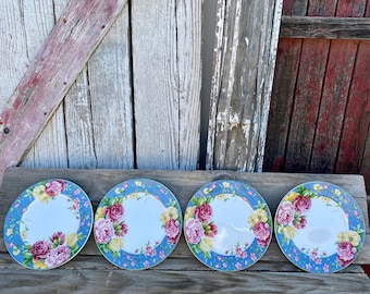 May include: Four floral dinner plates with a blue and white background and a colorful floral design. The plates are arranged in a row on a wooden surface.