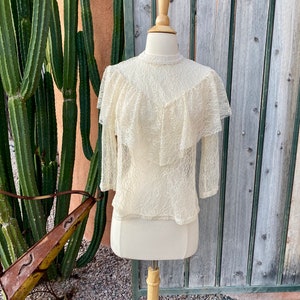 May include: A white lace top with a ruffled neckline and long sleeves. The top is on a mannequin against a wooden background.
