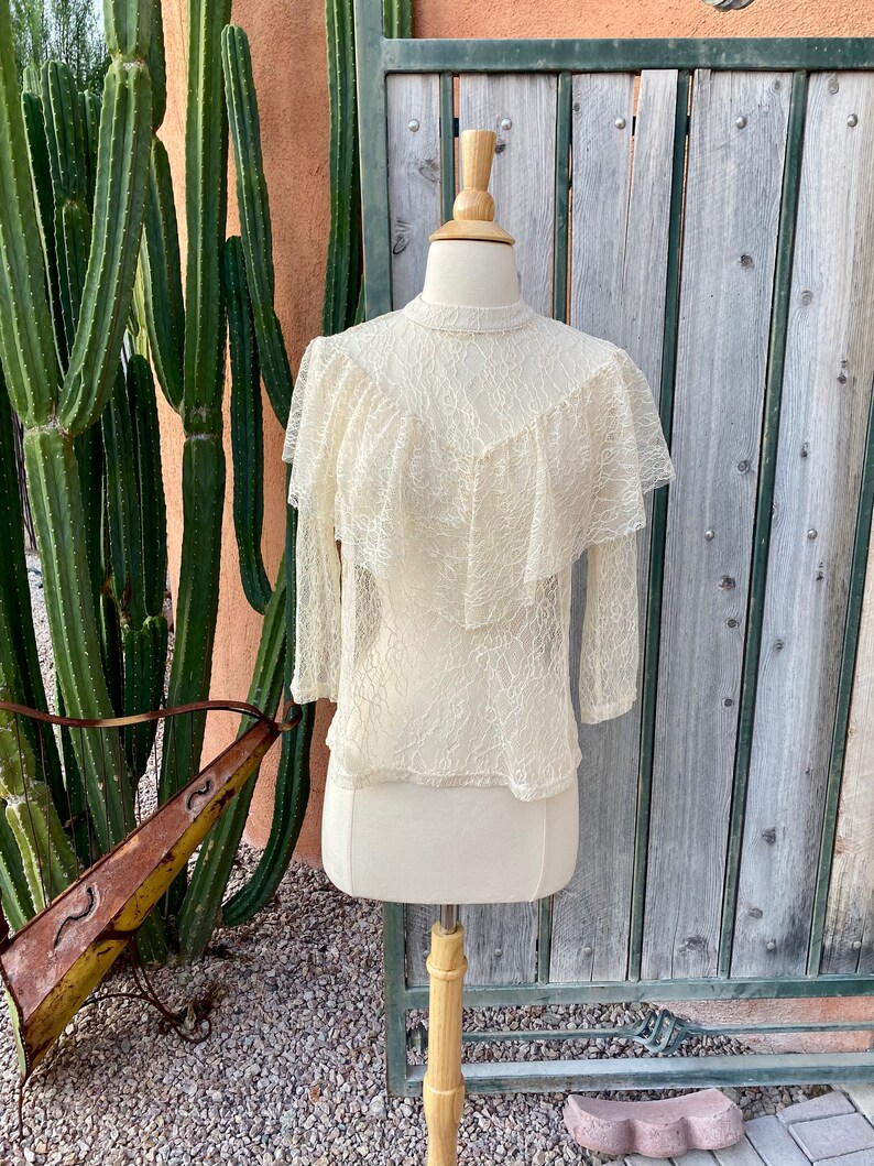 May include: A white lace top with a ruffled neckline and long sleeves. The top is on a mannequin against a wooden background.