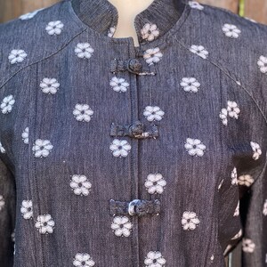May include: A black denim jacket with white embroidered flowers and a mandarin collar. The jacket has a series of decorative buttons down the front.