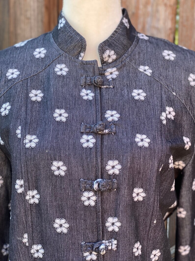 May include: A black denim jacket with white embroidered flowers and a mandarin collar. The jacket has a series of decorative buttons down the front.
