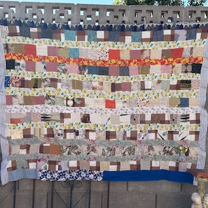 May include: A patchwork quilt with a variety of fabrics and patterns in shades of brown, white, and blue. The quilt is made up of squares and rectangles, and it has a fringed edge.