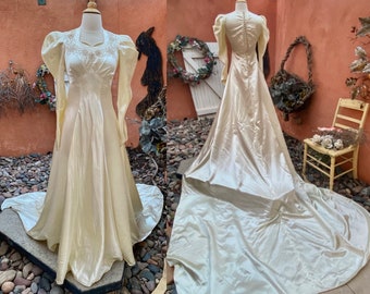 May include: A vintage ivory satin wedding dress with long sleeves and a fitted bodice. The dress has a full skirt and a train. The dress is on a mannequin and is displayed against a backdrop of a brick wall and greenery.