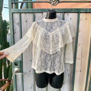 May include: A white lace top with a ruffled neckline and long sleeves. The top is sheer and has a delicate floral pattern.