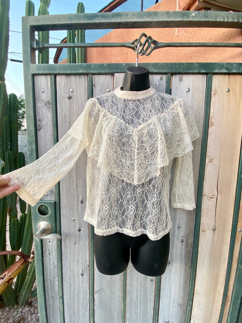 May include: A white lace top with a ruffled neckline and long sleeves. The top is sheer and has a delicate floral pattern.