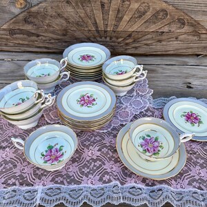 May include: A set of vintage teacups and saucers with floral designs. The cups and saucers are white with gold trim and feature pink and purple flowers. The set is arranged on a lace tablecloth.