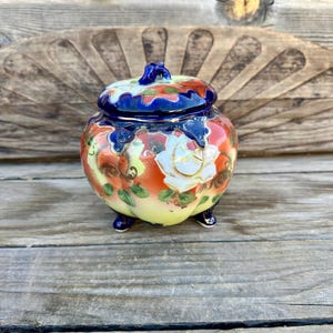 May include: A small, round, ceramic jar with a lid. The jar is decorated with a floral pattern in shades of red, white, yellow, and blue. The jar has three small feet.
