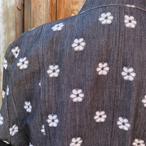 May include: A black denim jacket with a white floral pattern. The jacket has a collar and long sleeves.