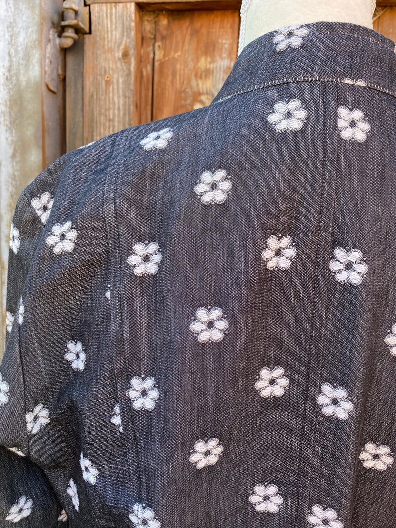 May include: A black denim jacket with a white floral pattern. The jacket has a collar and long sleeves.