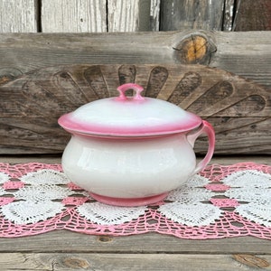 May include: A white ceramic chamber pot with a pink rim and lid. The pot has a single handle and is sitting on a pink and white crocheted doily.