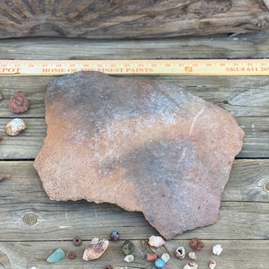 May include: A large, irregularly shaped, reddish-brown stone slab with a lighter, mottled surface. Small, colorful stones and beads are scattered around the slab on a wooden surface. A wooden ruler with text is visible.