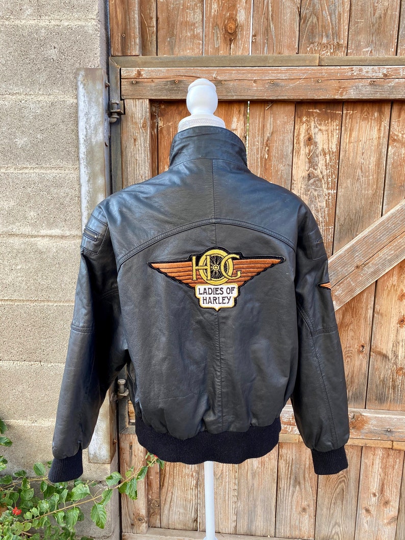Puede incluir: Chaqueta de cuero negra con un parche bordado en marr&oacute;n y dorado que dice "Ladies of Harley" y un logotipo alado con las letras "HOG".