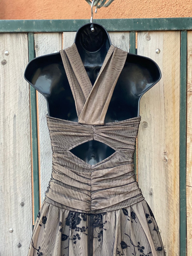 May include: A brown and black formal dress with a criss-cross halter neckline and a keyhole cutout in the back. The dress has a tulle skirt with black floral embroidery.