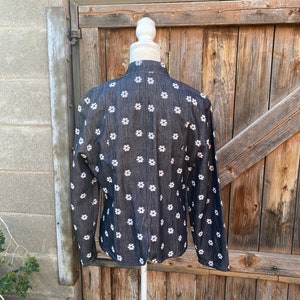 May include: A black denim jacket with a white floral pattern. The jacket is on a white mannequin and is displayed against a wooden background.