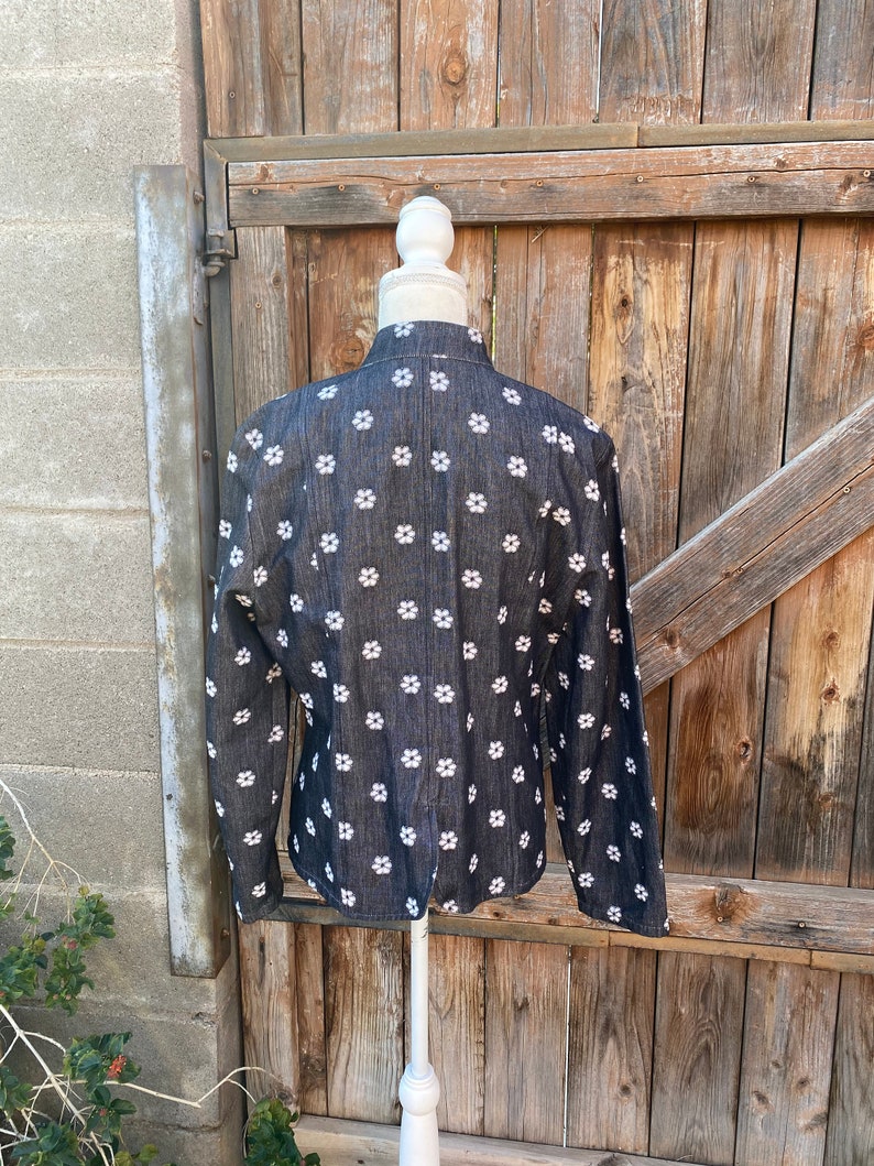 May include: A black denim jacket with a white floral pattern. The jacket is on a white mannequin and is displayed against a wooden background.