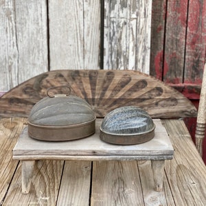 May include: Two vintage metal dome-shaped containers with lids, one with a handle, sitting on a rustic wooden stand. The stand has three legs and is on a wooden table.