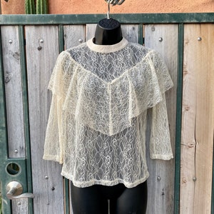 May include: A white lace top with a ruffled neckline and long sleeves. The top is sheer and has a delicate floral pattern.