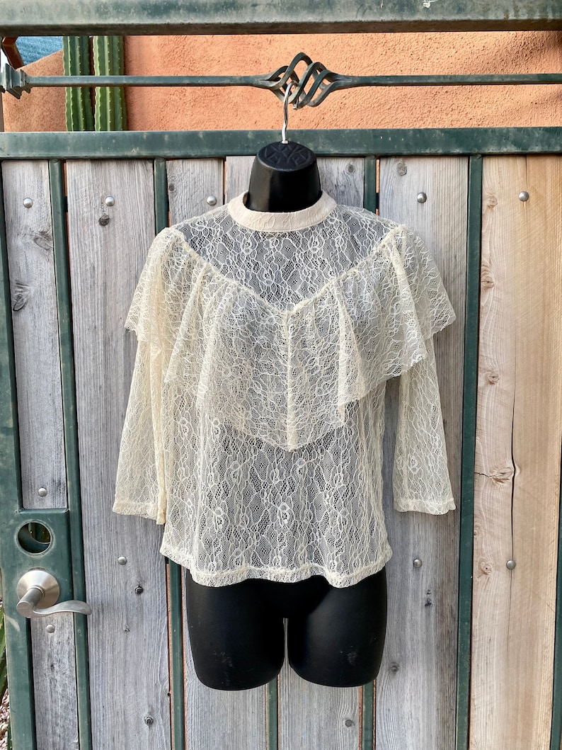 May include: A white lace top with a ruffled neckline and long sleeves. The top is sheer and has a delicate floral pattern.