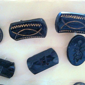 May include: Assortment of black glass buttons with various designs, including a cross, a floral pattern, and a geometric pattern. Some buttons have gold accents.
