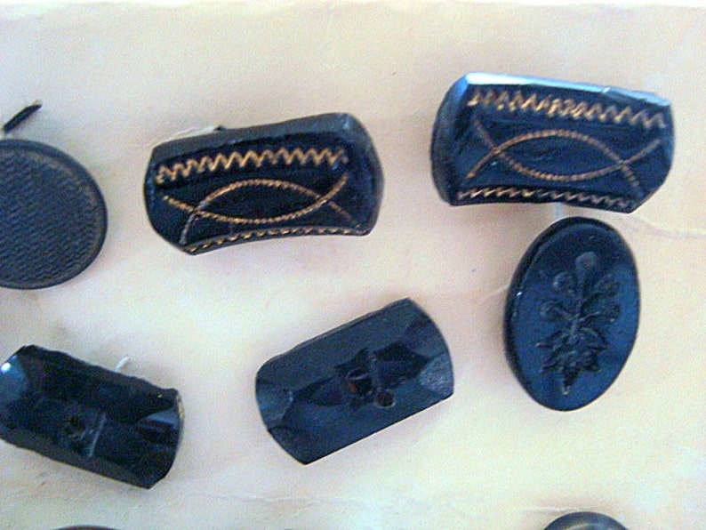 May include: Assortment of black glass buttons with various designs, including a cross, a floral pattern, and a geometric pattern. Some buttons have gold accents.