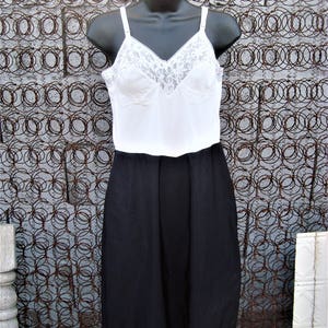 May include: A black slip with a lace trim. The slip is made of a smooth, silky fabric and has a fitted bodice. The slip is on a white mannequin against a rusty metal background.