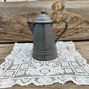 May include: A vintage gray enamelware coffee pot with a lid and handle. The pot sits on a white lace doily, with a rustic wooden background. The pot has a classic, old-fashioned design.