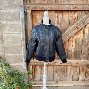 Puede incluir: Una chaqueta de cuero negra con cierre de cremallera y cuello y pu&ntilde;os de punto de canal&eacute;. La chaqueta tiene un parche en el hombro izquierdo y un bolsillo con cremallera en el pecho izquierdo.