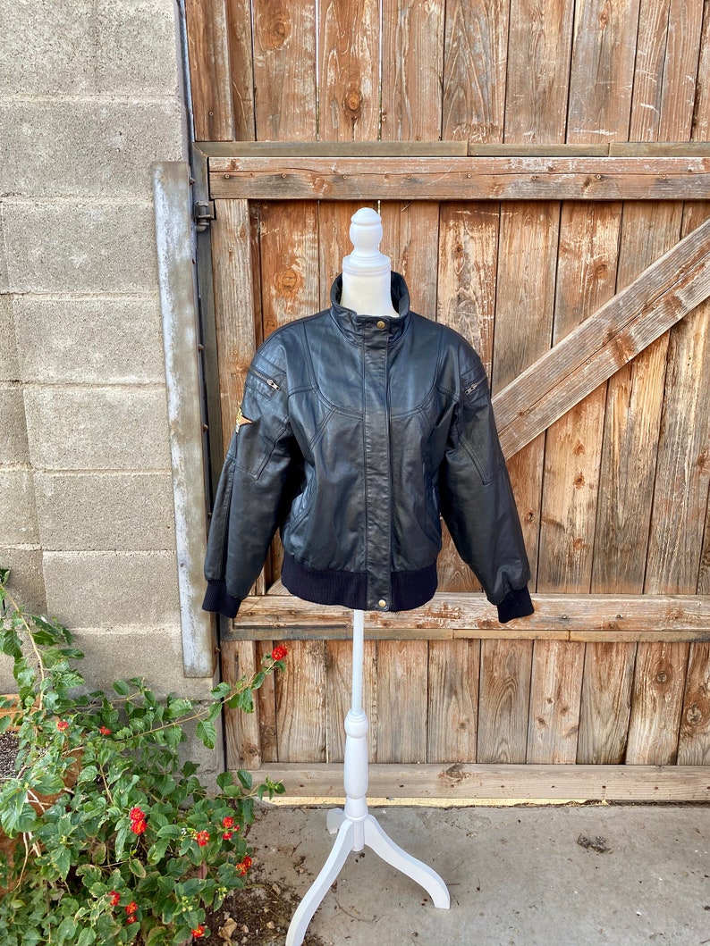 Puede incluir: Una chaqueta de cuero negra con cierre de cremallera y cuello y pu&ntilde;os de punto de canal&eacute;. La chaqueta tiene un parche en el hombro izquierdo y un bolsillo con cremallera en el pecho izquierdo.