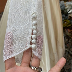 May include: Close-up of a white lace wedding dress sleeve with a row of pearl buttons.