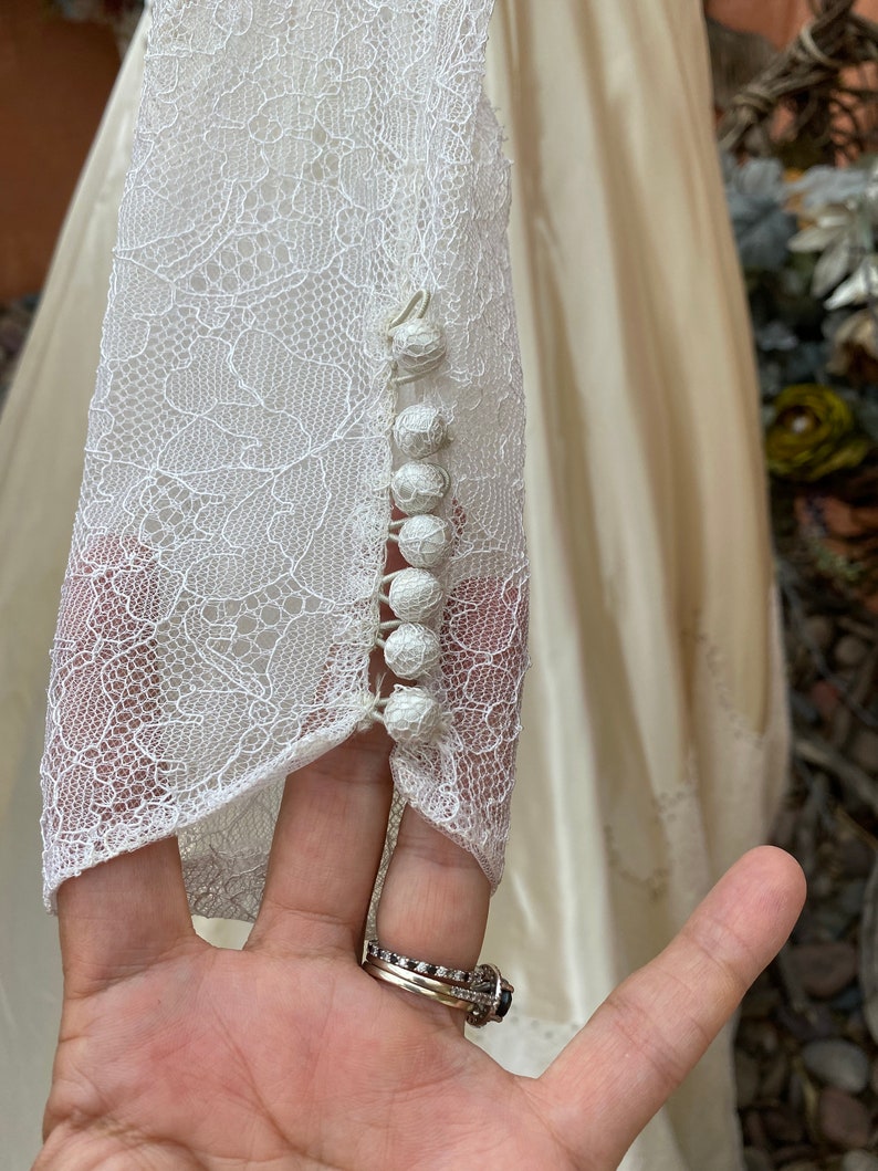 May include: Close-up of a white lace wedding dress sleeve with a row of pearl buttons.