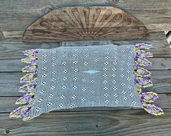 May include: A white, rectangular crocheted doily with a delicate openwork pattern. The edges are trimmed with yellow and purple floral designs. A wooden, sunburst-shaped decorative piece is in the background.