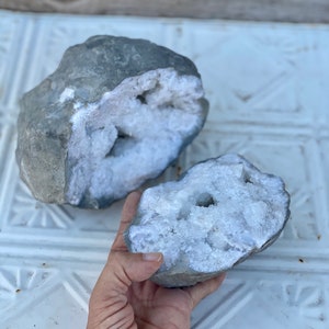 May include: Two gray rocks with white crystal formations inside. The rocks are broken open to reveal the crystals.