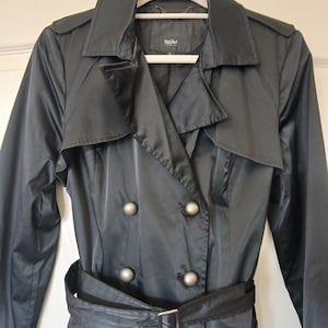 May include: A black trench coat with a double breasted closure and a belt. The coat has silver buttons and a label that says "Mango" on the inside.