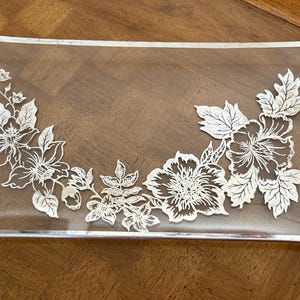 May include: A clear glass serving tray with a silver floral design. The design features a variety of flowers and leaves, all in a delicate, intricate style.