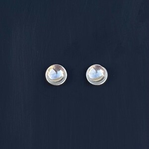 May include: A pair of silver stud earrings with a domed, reflective surface. The earrings have a simple, minimalist design. They are set against a dark blue background, which accentuates their bright, metallic appearance. These earrings are a classic jewellery accessory.