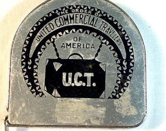 May include: A silver-toned metal tape measure case with a semi-circular top. The front features a black graphic with the text "THE ORDER OF UNITED COMMERCIAL TRAVELERS OF AMERICA" and "U.C.T." centered above a depiction of a vintage suitcase.