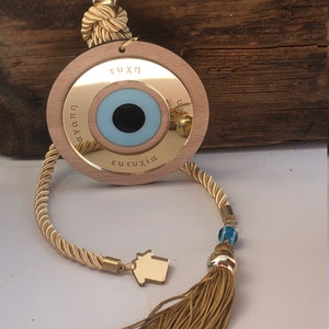 May include: A wooden evil eye charm with a blue eye and gold trim. The charm is attached to a gold tassel and a beige rope. The charm has the words "&tau;ύ&chi;&eta;", "&alpha;&gamma;ά&pi;&eta;", and "&epsilon;&upsilon;&tau;&upsilon;&chi;ί&alpha;" inscribed on it.