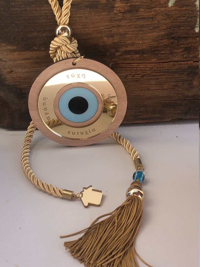 May include: A wooden evil eye charm with a blue eye and gold trim. The charm is attached to a gold tassel and a beige rope. The charm has the words "&tau;ύ&chi;&eta;", "&alpha;&gamma;ά&pi;&eta;", and "&epsilon;&upsilon;&tau;&upsilon;&chi;ί&alpha;" inscribed on it.