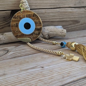 Op de afbeelding: Gouden boze oog charme met een blauw oog en een gouden kwast. De charme heeft de tekst "Tuti & Yianni" en "Agapi" erop gegraveerd.