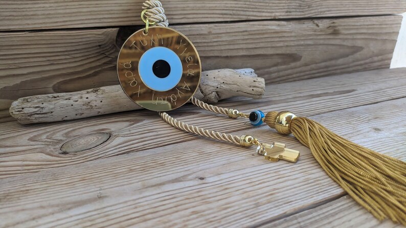 Op de afbeelding: Gouden boze oog charme met een blauw oog en een gouden kwast. De charme heeft de tekst "Tuti & Yianni" en "Agapi" erop gegraveerd.