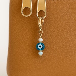Pu&ograve; includere: Primo piano di un astuccio in pelle marrone con cerniera dorata e un ciondolo decorativo. Il ciondolo presenta un design con perline a forma di occhio blu e bianco, aggiungendo un tocco di eleganza. L'astuccio &egrave; di un colore caldo e neutro.