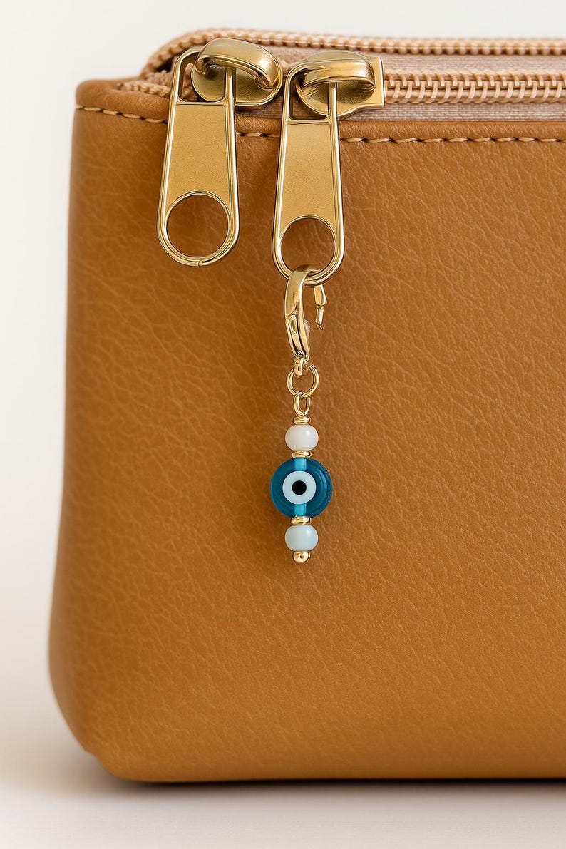 Pu&ograve; includere: Primo piano di un astuccio in pelle marrone con cerniera dorata e un ciondolo decorativo. Il ciondolo presenta un design con perline a forma di occhio blu e bianco, aggiungendo un tocco di eleganza. L'astuccio &egrave; di un colore caldo e neutro.