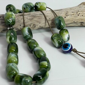 Op de afbeelding: Een groene en witte marmeren gebedskralen ketting met een blauwe kwade oog charme. De kralen zijn geregen op een bruin koord.